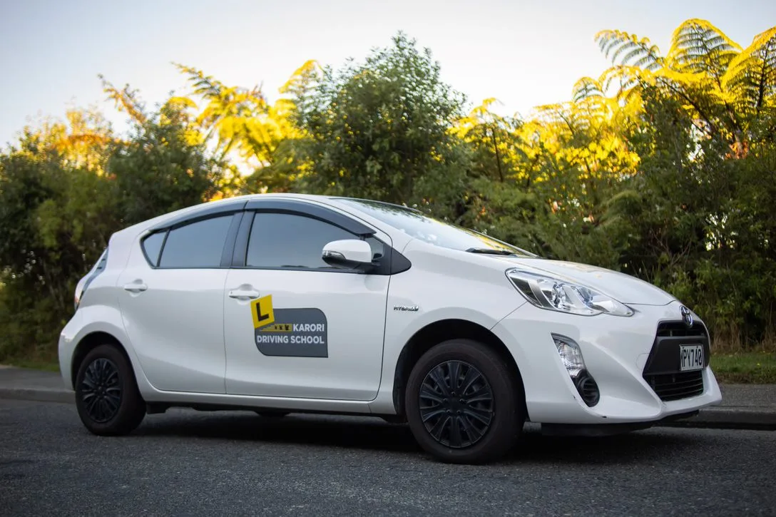 A white Toyota Aqua with Karori Driving School signage parked on the side of a road.